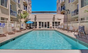 Une piscine rectangulaire à l’eau d’un bleu limpide, entourée de chaises longues et de parasols, avec un bâtiment doté de balcons et un panneau indiquant la présence d’un sauveteur en service en arrière-plan.