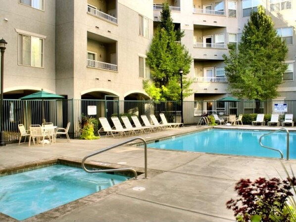 A swimming pool with lounge chairs and umbrellas, surrounded by a building with balconies and trees.