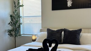 Un lit bien fait, agrémenté d’une serviette pliée en forme de cœur, une peinture abstraite en noir et blanc accrochée au mur, et une plante en pot placée près de la fenêtre.