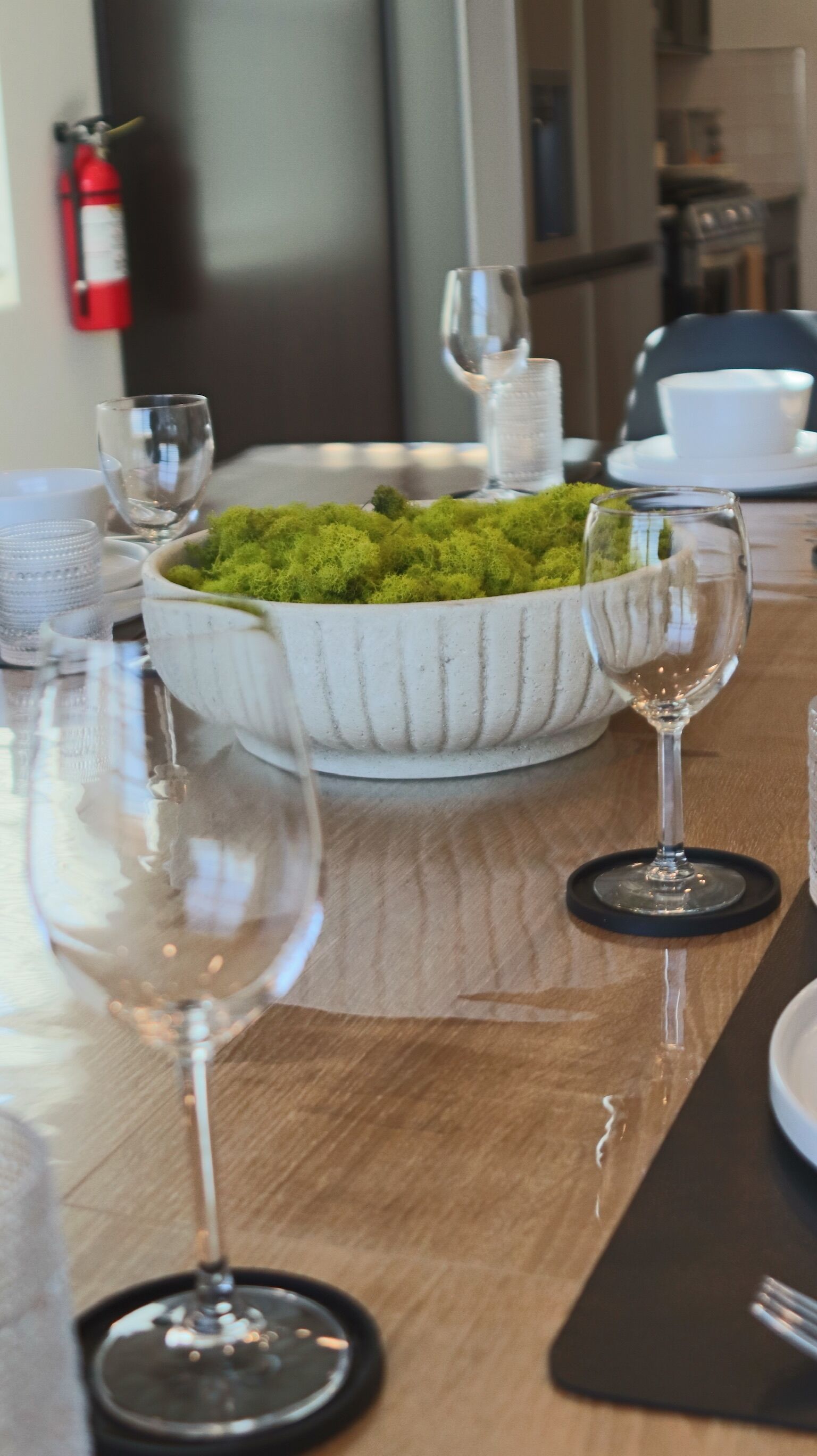 Una mesa de comedor con un bol blanco de musgo verde, dos copas de vino y un extintor de incendios al fondo.