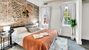 Ein Schlafzimmer mit Backsteinwand, einem Bett mit brauner Bettdecke, weißen Kissen und einem kleinen Nachttisch mit Lampe.