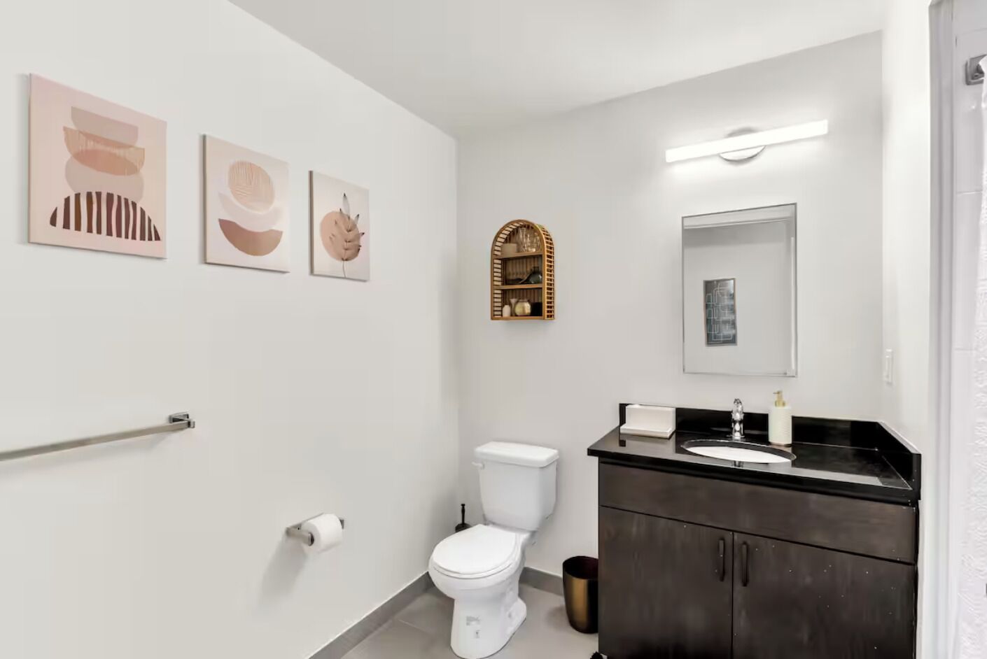 Un baño moderno con un inodoro blanco, un tocador negro con un espejo y tres obras de arte abstracto en la pared.