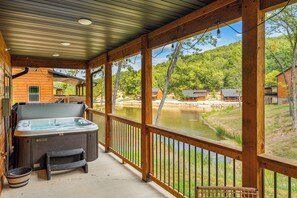 A wooden deck with a hot tub, a wooden chair, and a metal railing overlooking a lake and forested area. - Cozy New Cabin with Private Hot Tub, Free Local Attraction Tickets & Community Pools (Hollister)
