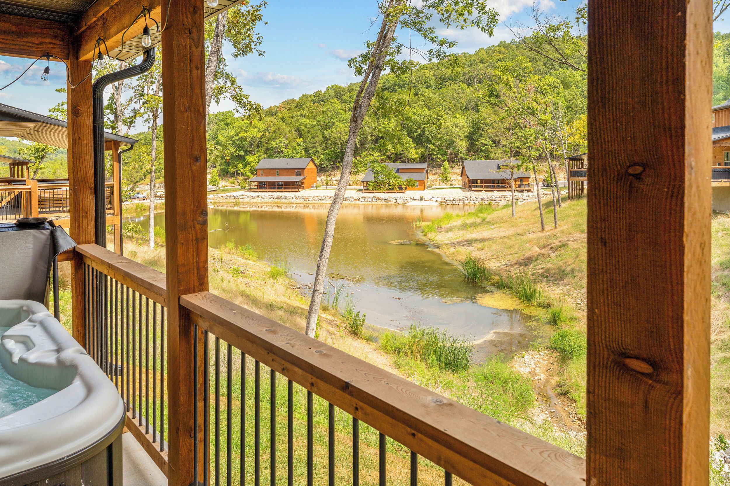 A wooden deck with a hot tub, overlooking a lake with cabins and trees.