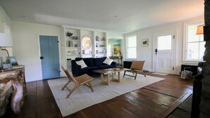 Una sala de estar con un sofá azul oscuro, una mesa de centro de madera y dos sillas de madera. Hay una estantería blanca llena de objetos decorativos y una puerta con un panel azul.