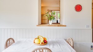 Un coin repas avec une table dressée pour deux personnes, un panier de fruits sur la table et une horloge au mur.