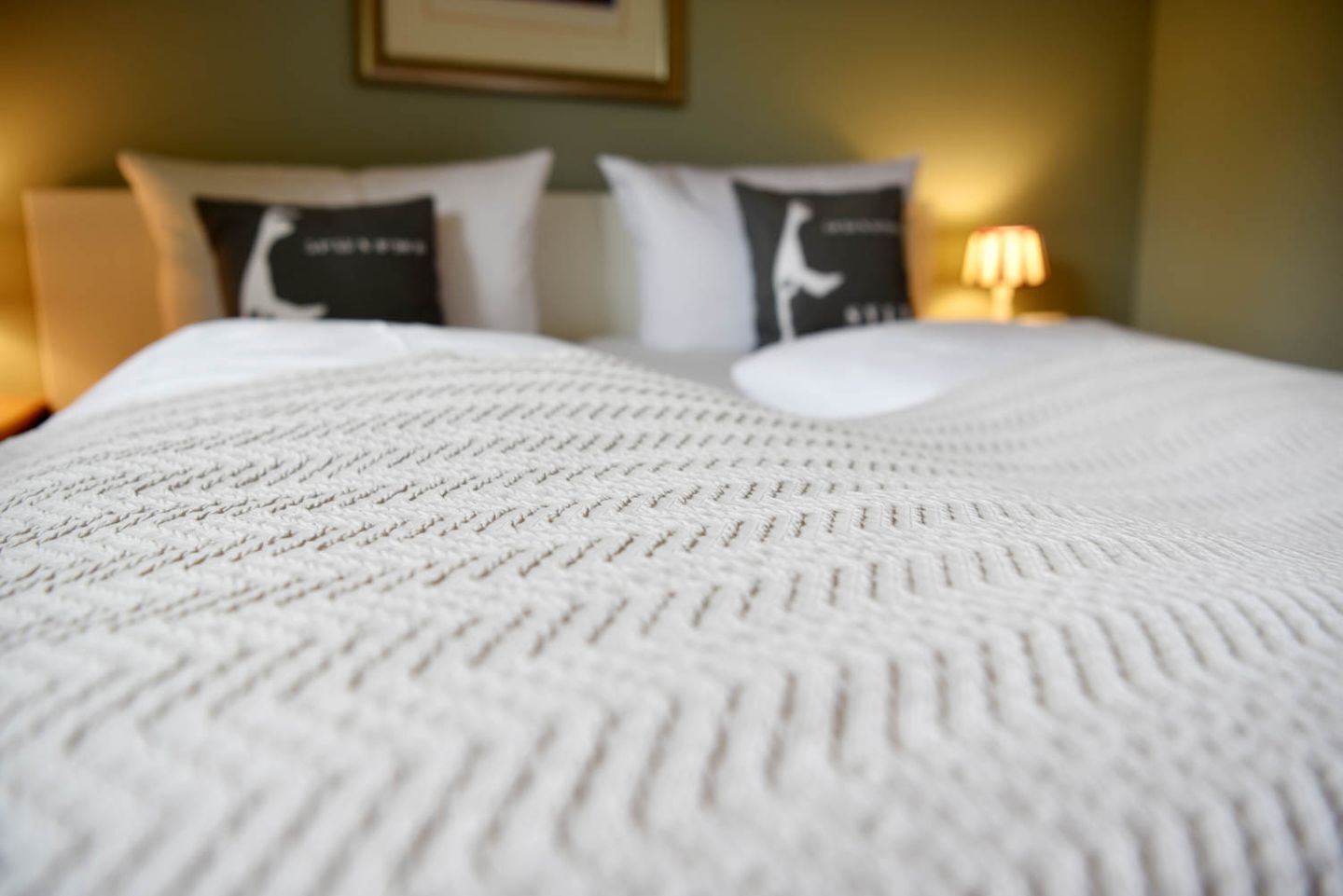 A bed with a textured white blanket, two pillows with black and white designs, and a wall-mounted picture frame.