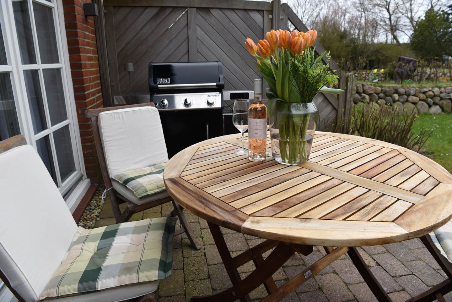 An outdoor patio with a round wooden table, a white cushioned chair, a grill, and a vase with flowers.