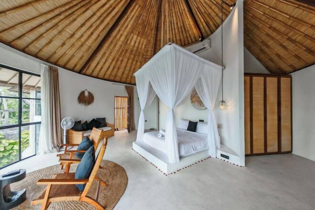 Une pièce dotée d’un toit de chaume, d’un lit à baldaquin, d’un coin salon avec des fauteuils et offrant une vue dégagée grâce à de grandes fenêtres.