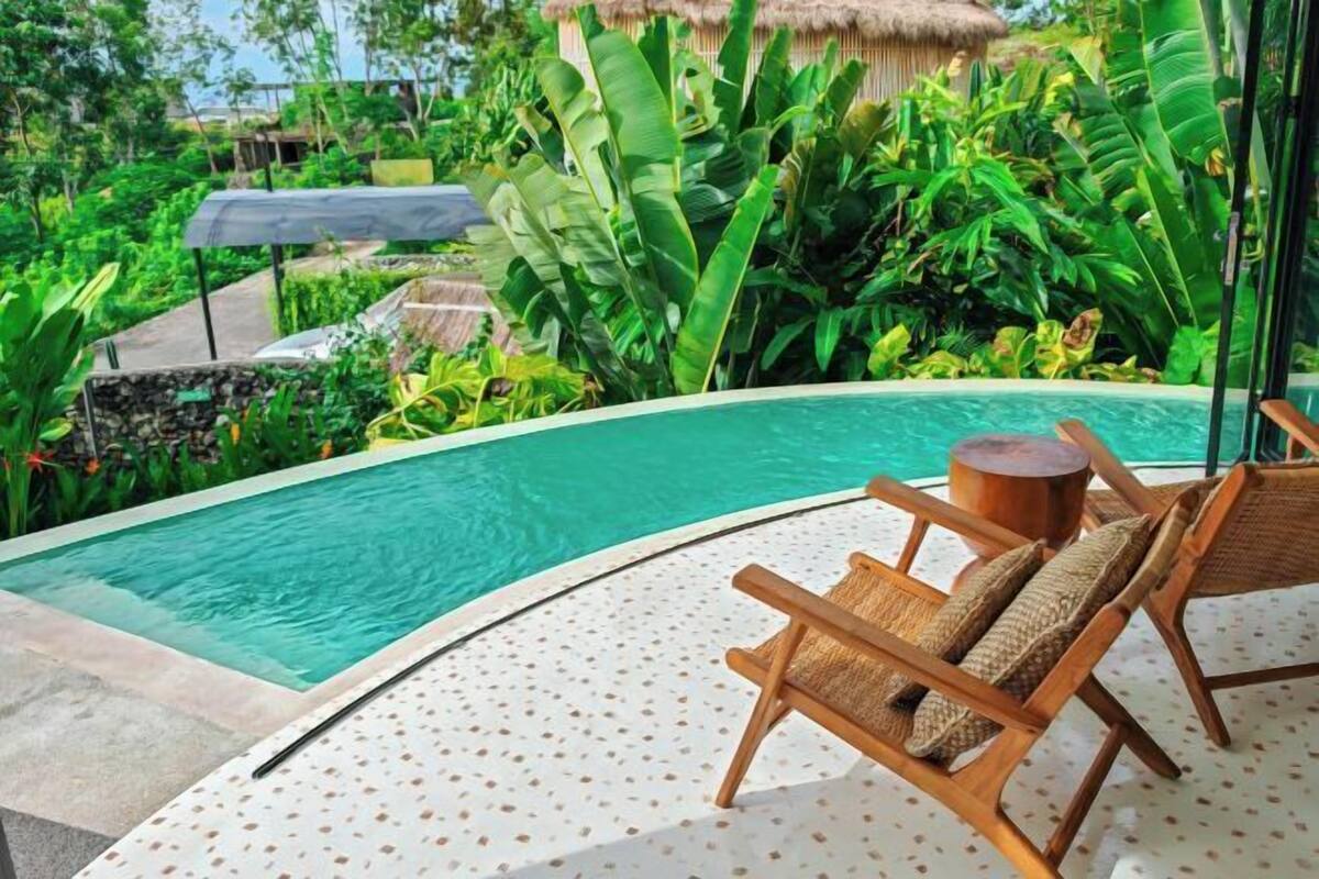Un espace aménagé au bord de la piscine, avec un fauteuil de relaxation, une table ronde en bois et une végétation tropicale luxuriante.