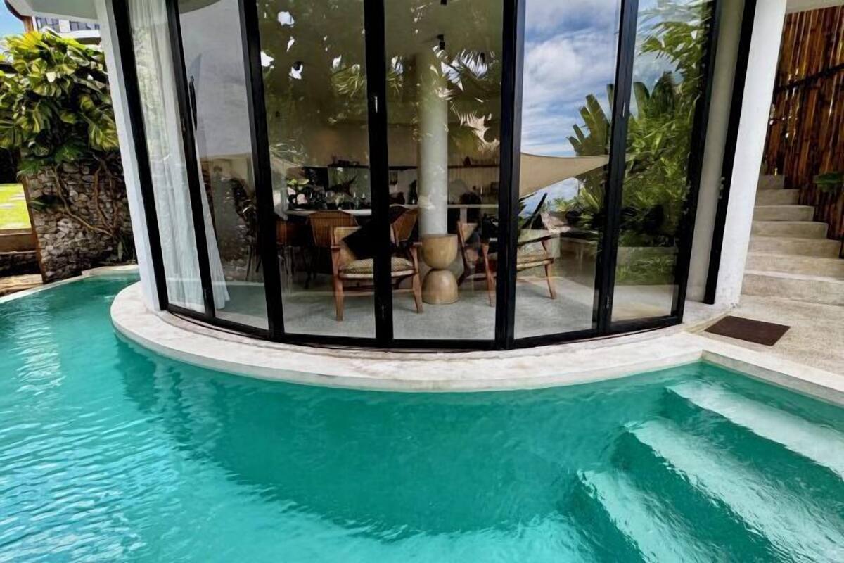 Un espace piscine protégé par une paroi en verre, un coin repas avec chaises et tables, et un escalier menant à un niveau supérieur.