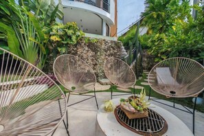 An outdoor seating area with wicker chairs and a round table, surrounded by lush greenery and a stone wall.