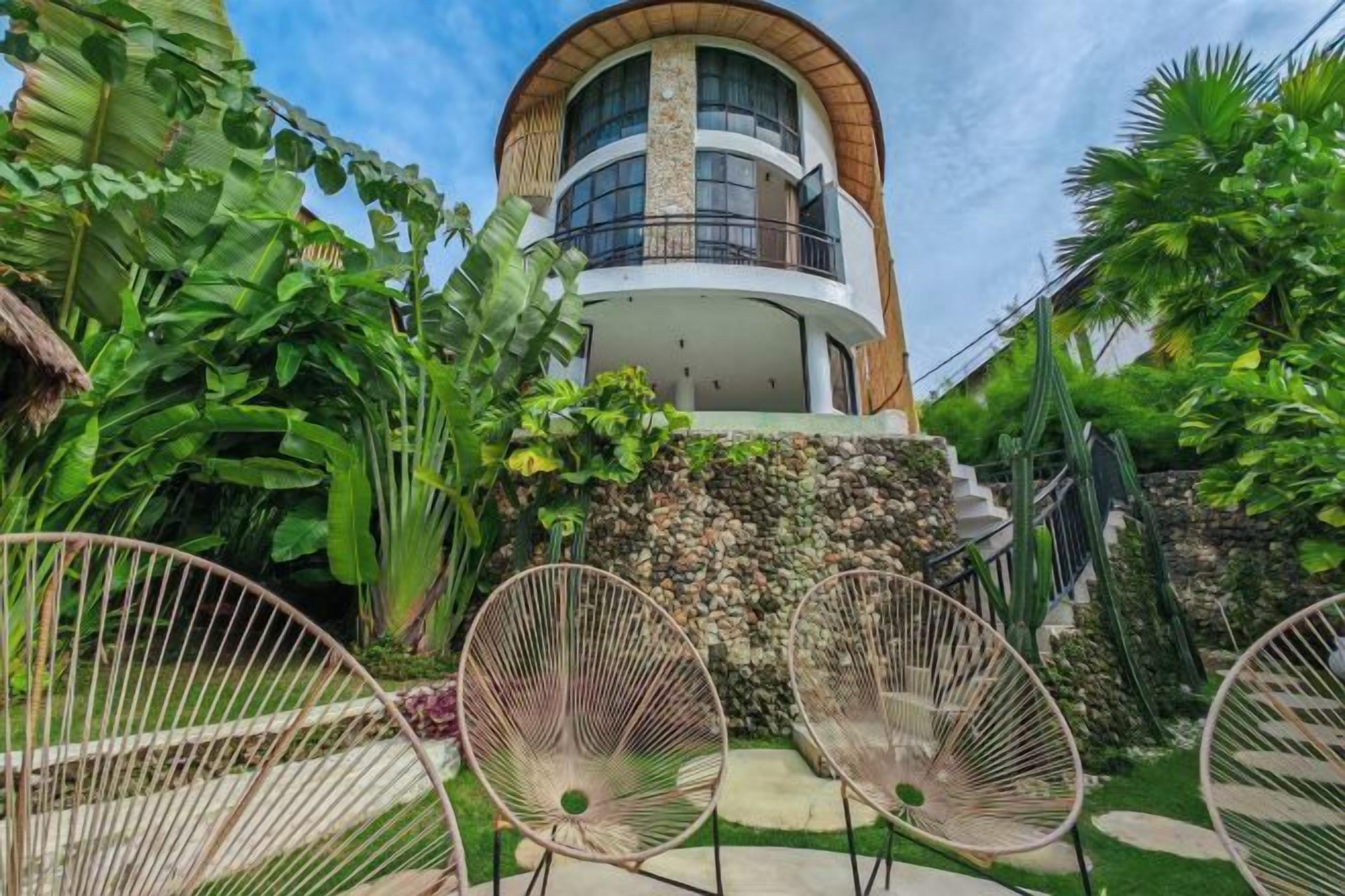 Een modern huis met een gebogen balkon, omringd door weelderige begroeiing en tuinmeubilair.
