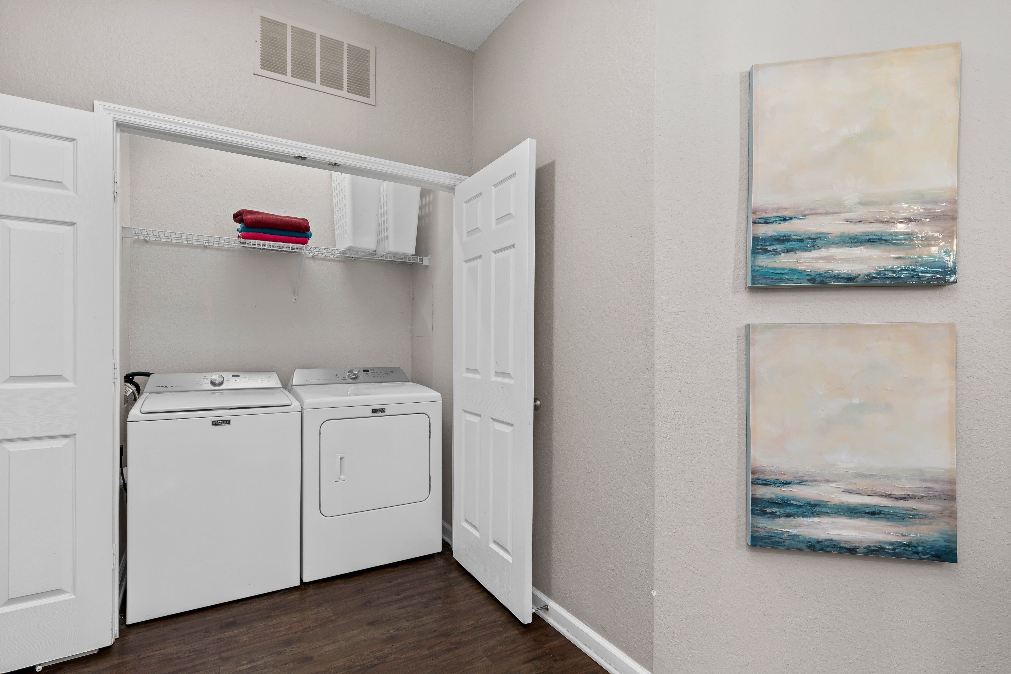 A laundry room with a washer and dryer, a shelf with folded towels, and two paintings on the wall.