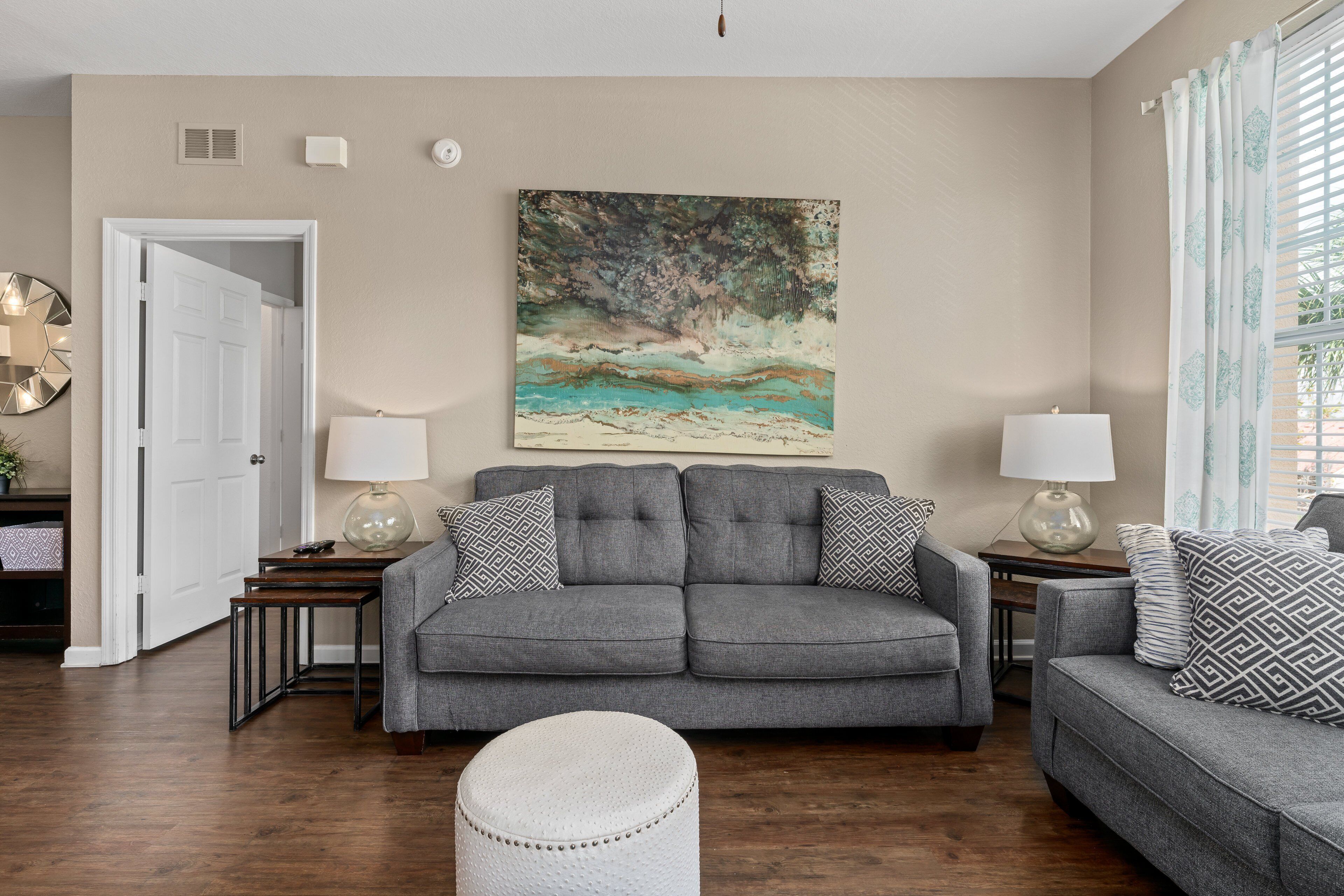 Un salon avec un canapé gris, deux tables d’appoint avec des lampes, un pouf blanc et un grand tableau abstrait accroché au mur.