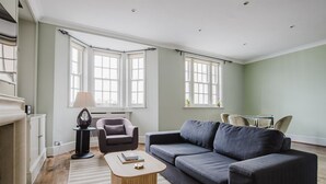 A modern living room with a grey sofa, a wooden coffee table, and a black armchair. There are two windows with white curtains, a radiator, and a small table with chairs.