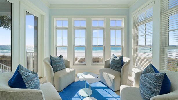 A sunlit room with large windows overlooking a beach, featuring two white armchairs with patterned cushions and a small white table. - Sea Glass - Huge Ocean Front Home with Gulf Views, Private Pool & Direct Beach Access (Miramar Beach)