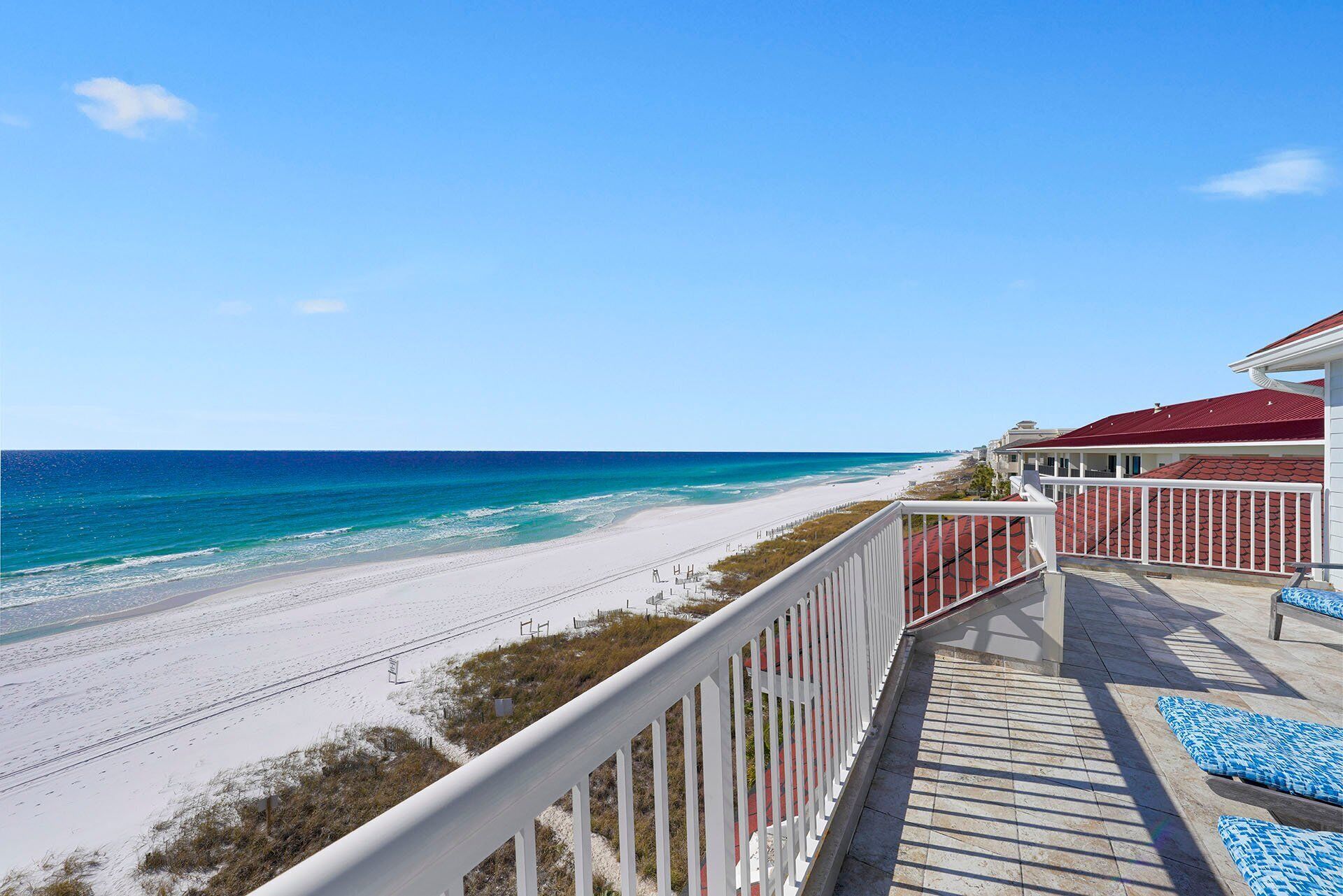 A beach view from a balcony with a railing, a pool area with blue patterned lounge chairs, and a building with a red roof.