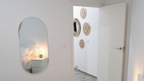 A hotel room with a bed, a round mirror, a bedside table with a lamp, and a door leading to another room.