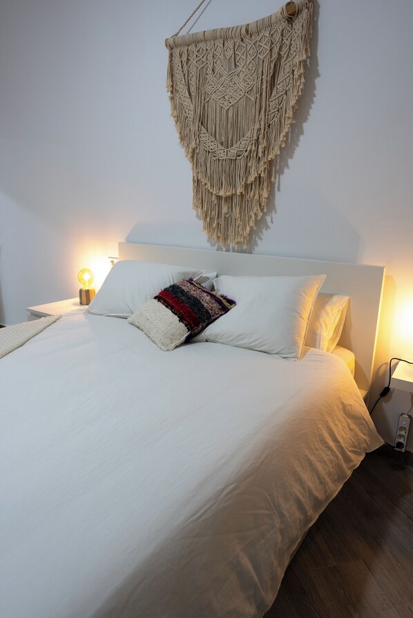 A neatly made bed with white linens and a colorful throw pillow, a macrame wall hanging, and bedside lamps.