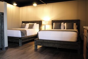 Two wooden beds with white linens and pillows, a nightstand, and a floor lamp in a room with wooden flooring and white walls. - 4th Street Lofts (Bemidji)