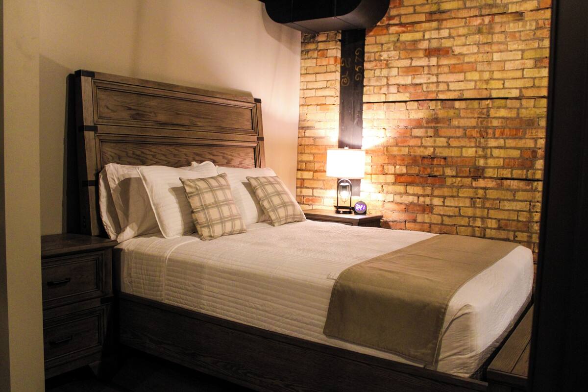 A neatly made bed with a wooden headboard, a nightstand with a lamp, and a brick-patterned wall.