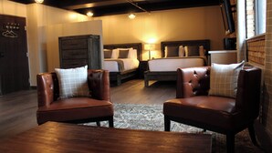 A hotel room with two leather armchairs, a wooden coffee table, a large bed with a headboard, a dresser, and a television on a stand. - 4th Street Lofts (Bemidji)
