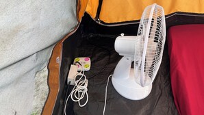 Ein Zelt mit einem weißen Ventilator, einer Steckleiste und einer roten Decke.