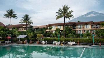 A swimming pool with lounge chairs and umbrellas, surrounded by palm trees and hotel buildings with a mountain in the background.