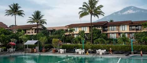 A swimming pool with lounge chairs and umbrellas, surrounded by palm trees and hotel buildings with a mountain in the background.