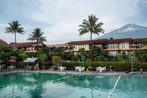 A swimming pool with lounge chairs and umbrellas, surrounded by palm trees and hotel buildings with a mountain in the background. - Grage Resort Sangkan (Cigandamekar)