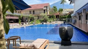 A swimming pool with lounge chairs and umbrellas, surrounded by buildings and palm trees. - Grage Resort Sangkan (Cigandamekar)