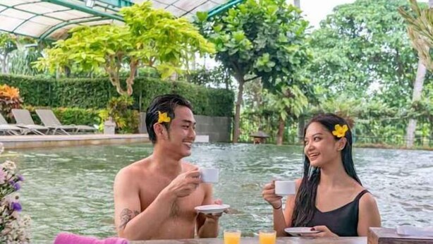 A couple dining by a pool with a glass table, surrounded by lush greenery and a covered area. - Grage Resort Sangkan (Cigandamekar)