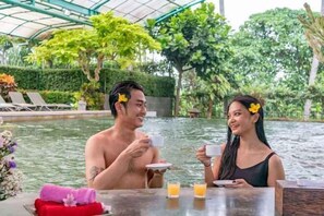 A couple dining by a pool with a glass table, surrounded by lush greenery and a covered area. - Grage Resort Sangkan (Cigandamekar)