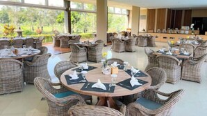 A spacious outdoor dining area with wicker furniture and round tables, surrounded by large windows offering a view of greenery. - Grage Resort Sangkan (Cigandamekar)