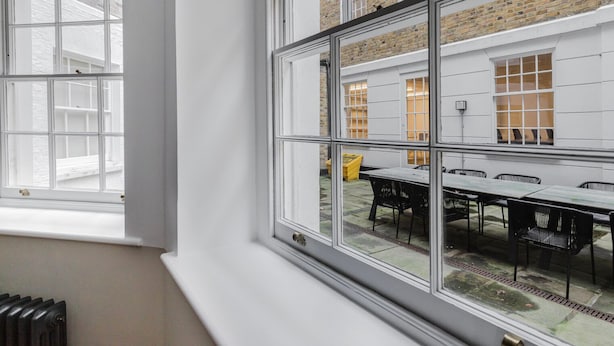 Ein Zimmer mit großem Fenster, weißen Wänden und Blick auf eine Terrasse mit Tisch und Stühlen.