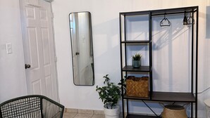 Una habitación con puerta, una silla, una planta en maceta, un espejo y un estante negro con cestas.