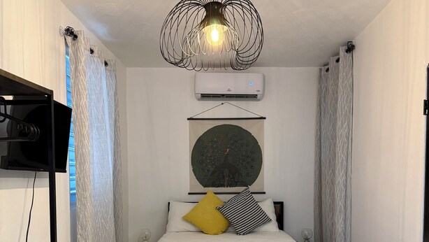 A bedroom with a bed, a wall-mounted air conditioner, a decorative light fixture, and patterned curtains. - Cozy_Studio 3@San Juan / Water Tank (San Juan)