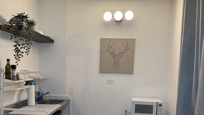 A compact kitchen with a microwave, sink, and a chair. There is a shelf with kitchen items and a wall-mounted light fixture. - Cozy_Studio 3@San Juan / Water Tank (San Juan)