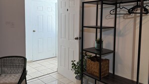 Un pasillo con una puerta blanca, un estante de metal negro con cestas y una silla de mimbre.