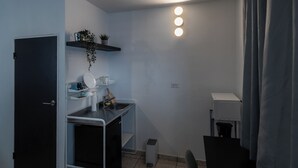A compact kitchen with a black door, a small countertop, and a sink. There is a shelf above the countertop holding dishes and a potted plant. A chair is positioned near the sink, and a curtain is partially visible on the right. - Cozy_Studio 3@San Juan / Water Tank (San Juan)