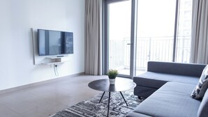 Een moderne woonkamer met een flatscreen televisie aan de muur, een grijze bank, een klein rond salontafeltje met een plant en een kamerbreed tapijt met een patroon.