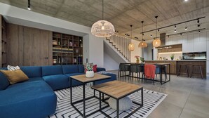 Ein modernes Wohnzimmer mit einem blauen Sofa, Holztischen, einer Treppe und einer Küchenzeile.