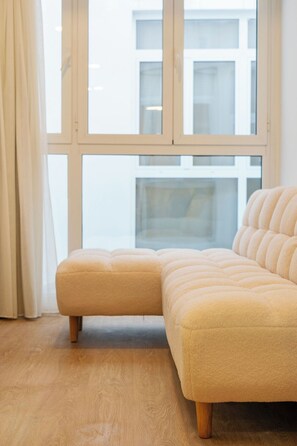 A corner sofa with a light-colored, tufted design, positioned in front of a large window with a view of a building. - VLC HOST- Molinell 3 (Valencia)