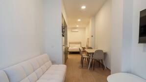 A modern hotel room with a sofa, a bed, a dining table with chairs, and a TV mounted on the wall.