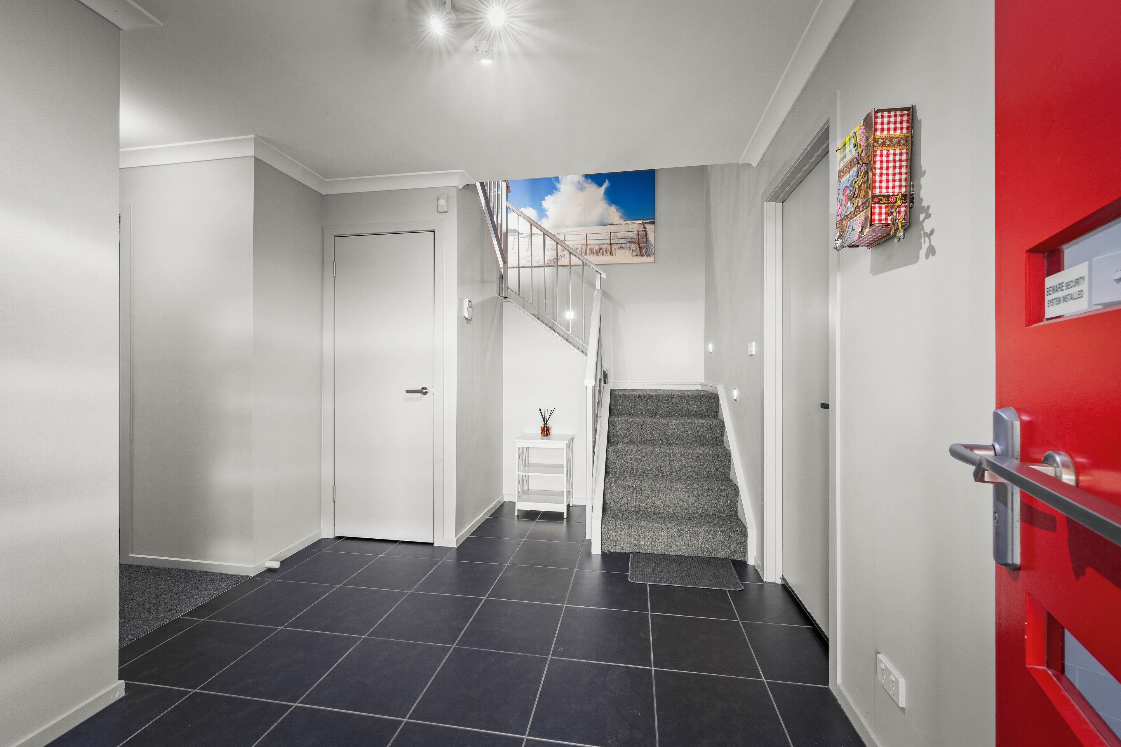 A modern hallway with a grey carpeted staircase, white walls, and a red door.