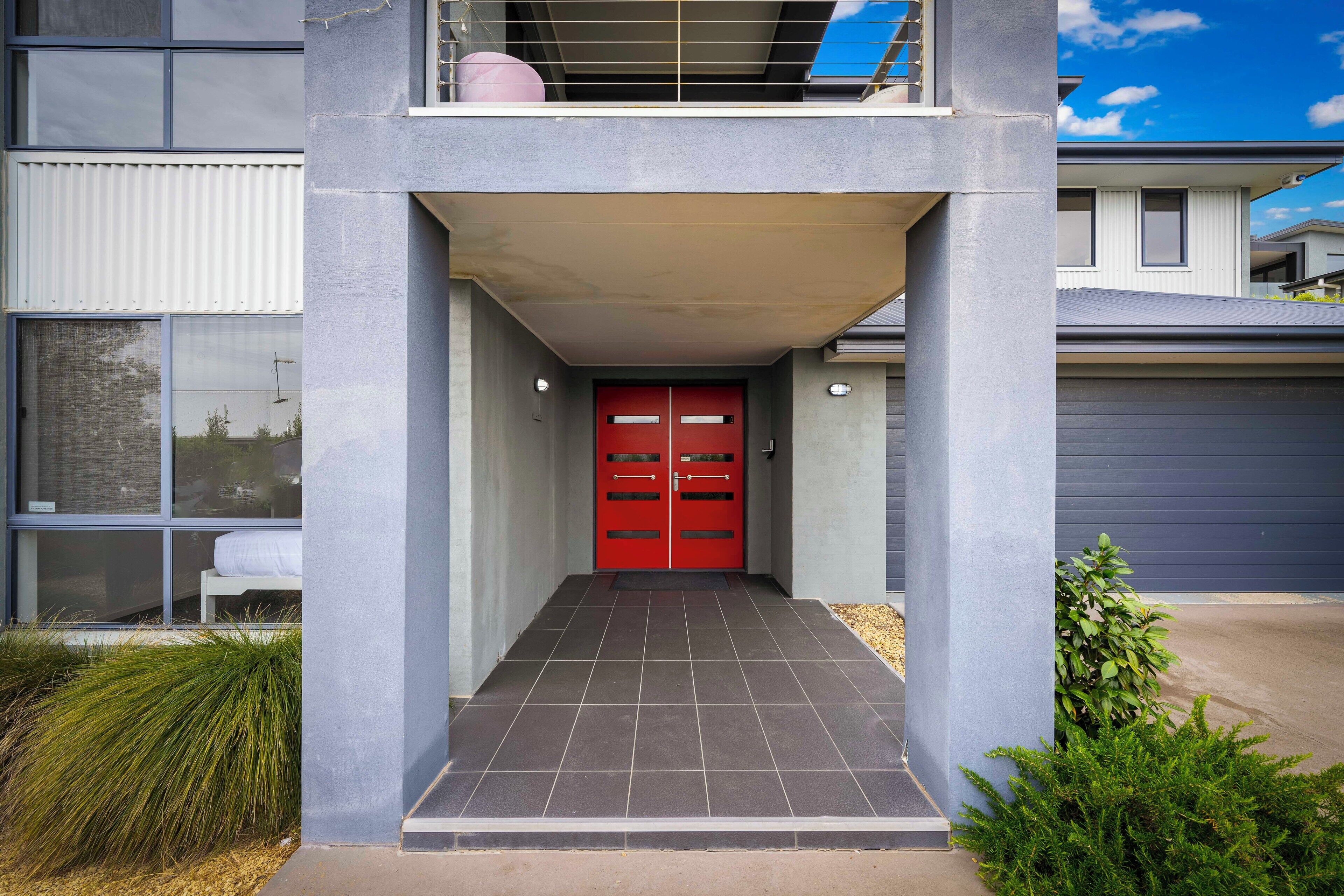 Un edificio moderno de dos pisos con una puerta roja, fachada gris y un acceso cubierto.