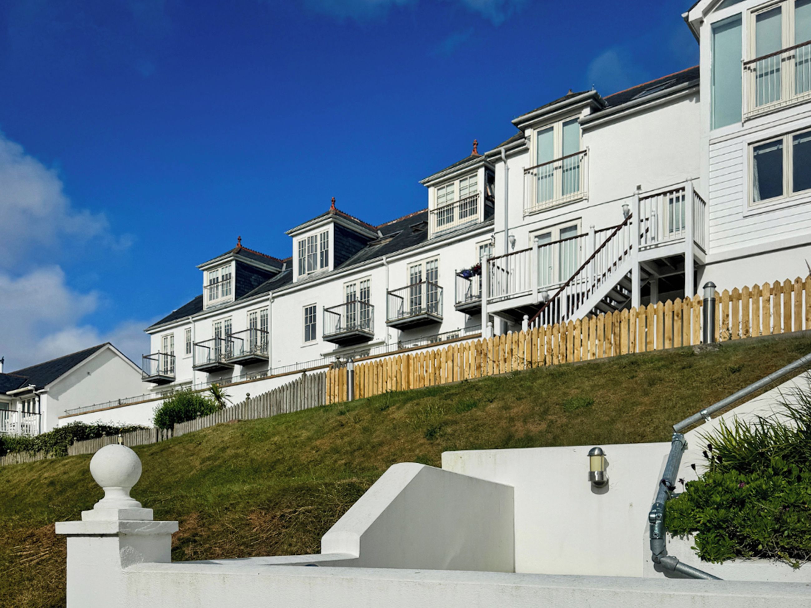 A row of white houses with balconies, a wooden fence, and a grassy hill.