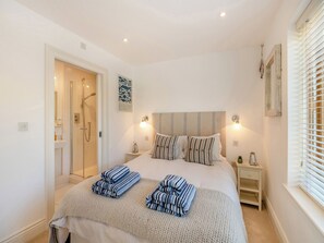 A bedroom with a bed, bedside tables, a nightstand, a window with blinds, a door leading to a bathroom, and a wall-mounted light fixture. - 2 bedroom accommodation in Hallsands (Hallsands)