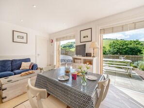 A cozy living room with a blue sofa, a dining table set for two, and a TV on a shelf. - 2 bedroom accommodation in Hallsands (Hallsands)
